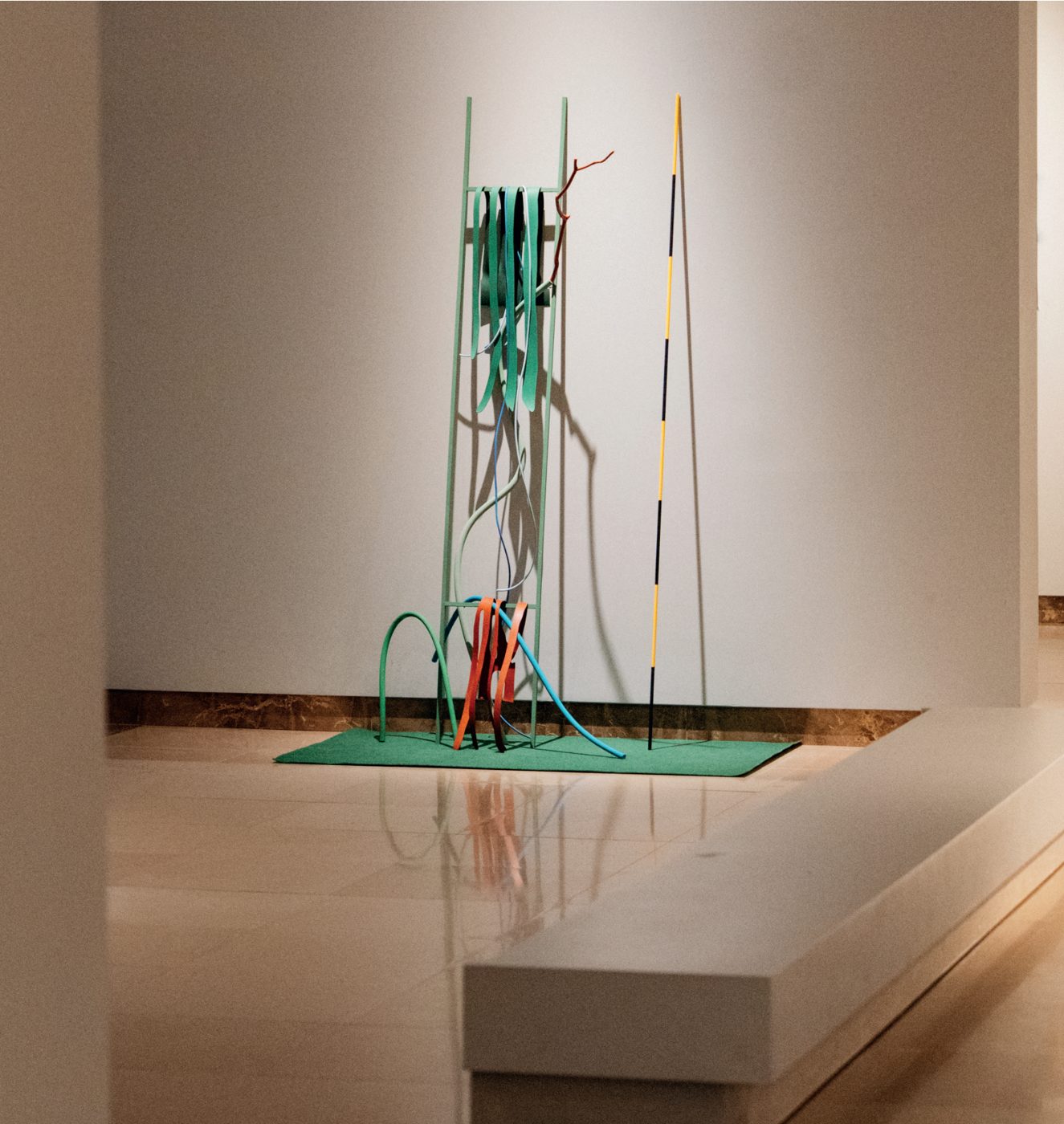 Escultura numa sala do museu: estrutura vertical verde com tiras e tubos de plástico coloridos, sobre um tapete de relva sintética.
